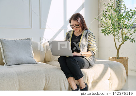 Portrait of middle aged woman using laptop sitting on sofa at home Portrait of middle aged woman using laptop sitting on sofa at home 142576119