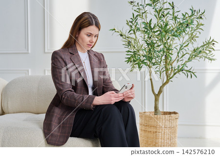 Young surprised woman sitting on couch in office using smartphone Young surprised woman sitting on couch in office using smartphone 142576210