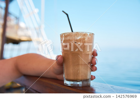 Close-up of a hand holding a refreshing iced frothy coffee drink by the sea Close-up of a hand holding a refreshing iced frothy coffee drink by the sea 142576969