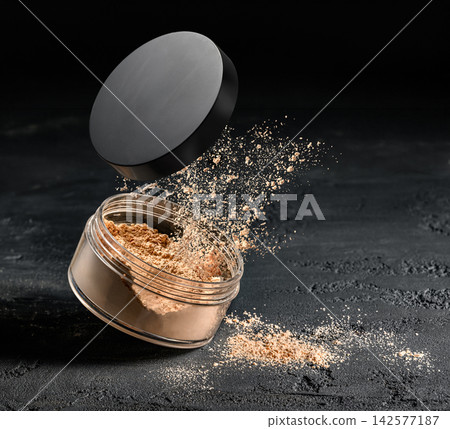 Close-up open loose face powder jar with puff releasing fine cosmetic powder cloud over dark textured tabletop 142577187