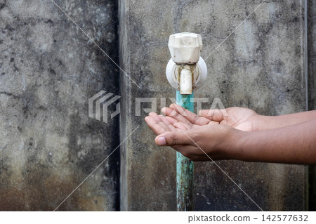 Human hands cupped under dry plastic water tap attached to weathered wall. Depicting water scarcity concept, global drought crisis, and resource shortage. Human hands cupped under dry plastic water tap attached to weathered wall. Depicting water scarcity concept, global drought crisis, and resource shortage. 142577632