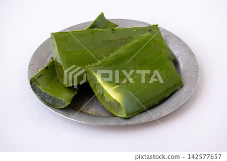 Fresh green sliced aloe vera leaves placed on metal plate over white background. Natural organic succulent plant prepared for skincare and herbal treatment. Fresh green sliced aloe vera leaves placed on metal plate over white background. Natural organic succulent plant prepared for skincare and herbal treatment. 142577657