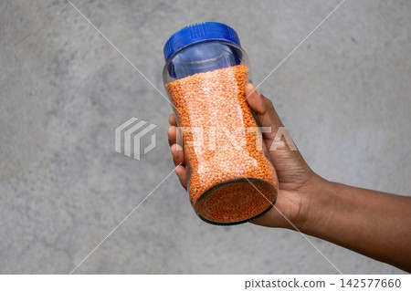 Hand holding transparent glass jar filled with red split lentils against rustic wall. Raw masoor dal stored in container for kitchen household pantry. Hand holding transparent glass jar filled with red split lentils against rustic wall. Raw masoor dal stored in container for kitchen household pantry. 142577660