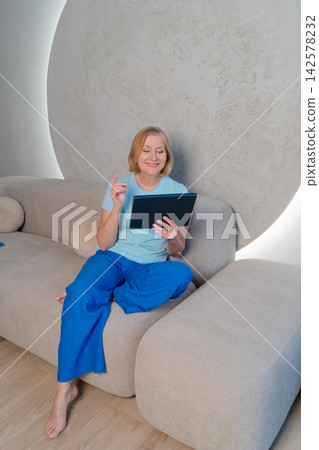 Senior lady with tablet smiling. Older Caucasian woman enjoys video chat. Elderly Caucasian female confidently interacts via video chat in cozy living room environment 142578232