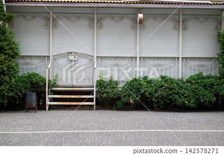 Closed white outdoor terrace with shutters, wrought iron gate, and green bushes Closed white outdoor terrace with shutters, wrought iron gate, and green bushes 142578271