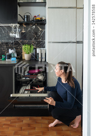 Woman Kneeling to Load the Dishwasher. A young woman kneels to load dishes into an open dishwasher in a modern home kitchen, doing daily household chores, a candid real-life lifestyle moment of housew 142578588