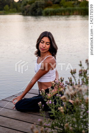 Woman in Seated Pose in a Summer Park. A young Hispanic woman practises a seated pose by the tranquil lake in a summer park at dusk, connecting body and mind in nature under a golden summer sky. 142578599