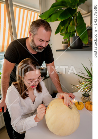 Father and Daughter Drawing on Pumpkin Together for Halloween at Home. Her father stands behind a daughter as she draws on a pale pumpkin at a festive table with mini pumpkins and a candle. A warm aut Father and Daughter Drawing on Pumpkin Together for Halloween at Home. Her father stands behind a daughter as she draws on a pale pumpkin at a festive table with mini pumpkins and a candle. A warm aut 142578680