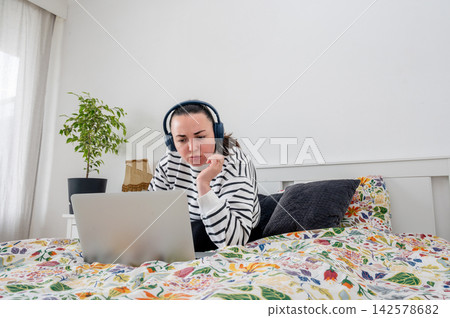 Woman studying on laptop lying in bed with headphones. Woman studying online material on laptop lying in bed with wireless headphones in white bedroom, e-learning from home and flexible education sche 142578682