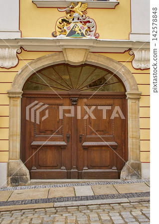 Large wooden double door with arched window and decorative coat of arms on building facade Large wooden double door with arched window and decorative coat of arms on building facade 142578848