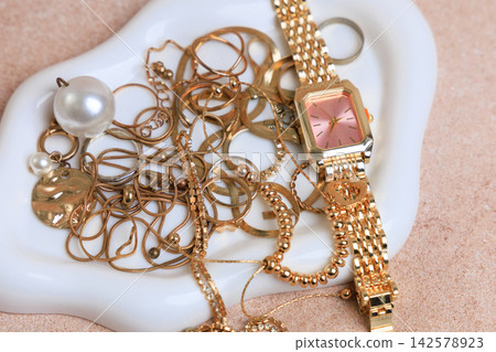 Various gold jewelry pieces and a watch arranged on a decorative plate in a well lit space during daytime Various gold jewelry pieces and a watch arranged on a decorative plate in a well lit space during daytime 142578923