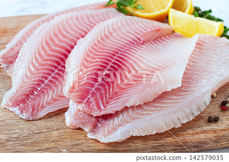Fresh Tilapia fillet, on a wooden chopping board, with lemon and herbs, ready to cook in the kitchen, no people Fresh Tilapia fillet, on a wooden chopping board, with lemon and herbs, ready to cook in the kitchen, no people 142579035