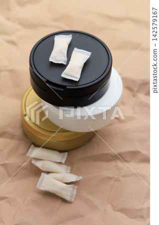 Different colors of snus pouches show nicotine pads on top of sealed containers placed on crumpled paper in a bright environment Different colors of snus pouches show nicotine pads on top of sealed containers placed on crumpled paper in a bright environment 142579167