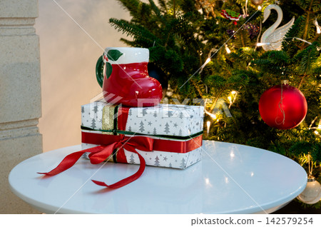 Red Santa Boot Mug on Gift with Red Bow by Christmas Tree. A glossy red Santa boot mug rests on a geometric gift with a red bow on a white table before a glowing Christmas tree with a red bauble. Warm Red Santa Boot Mug on Gift with Red Bow by Christmas Tree. A glossy red Santa boot mug rests on a geometric gift with a red bow on a white table before a glowing Christmas tree with a red bauble. Warm 142579254