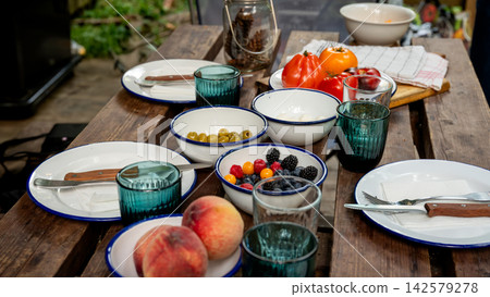 Al Fresco Picnic Table with Fresh Summer Produce in Garden. Rustic wooden picnic table set with white blue-rimmed enamel plates and bowls of olives, berries, tomatoes and peaches with blue glasses Al Fresco Picnic Table with Fresh Summer Produce in Garden. Rustic wooden picnic table set with white blue-rimmed enamel plates and bowls of olives, berries, tomatoes and peaches with blue glasses 142579278