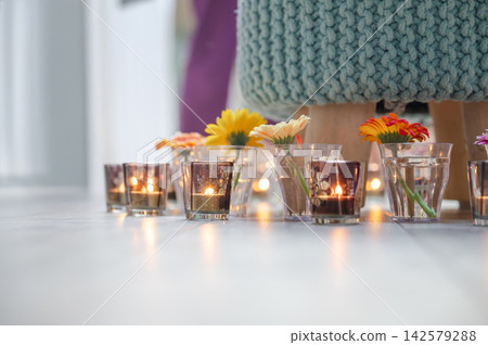 Tea Lights and Gerbera Flowers in Votives, Low Angle View. Low angle view of glowing tea light candles in amber glass votives alternating with colorful gerbera flowers on light wooden floor. Turquoise Tea Lights and Gerbera Flowers in Votives, Low Angle View. Low angle view of glowing tea light candles in amber glass votives alternating with colorful gerbera flowers on light wooden floor. Turquoise 142579288