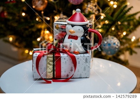 Penguin Mug on Festive Gift Box in Front of Christmas Tree. A novelty penguin mug in a Santa hat rests on a white gift box tied with a red bow before a glowing Christmas tree. Warm bokeh lights Penguin Mug on Festive Gift Box in Front of Christmas Tree. A novelty penguin mug in a Santa hat rests on a white gift box tied with a red bow before a glowing Christmas tree. Warm bokeh lights 142579289