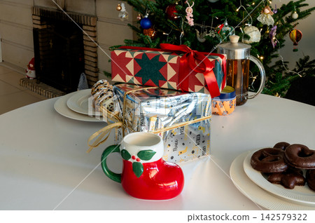 Boot Mug Gifts and French Press Tea on Christmas Table. A red Santa boot mug, festive wrapped gifts, a glass French press of tea and chocolate cookies on a white table before a glowing Christmas tree. Boot Mug Gifts and French Press Tea on Christmas Table. A red Santa boot mug, festive wrapped gifts, a glass French press of tea and chocolate cookies on a white table before a glowing Christmas tree. 142579322