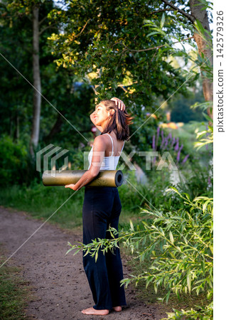Hispanic Woman in Woman Carrying a Yoga Mat at Golden Hour. A young Hispanic woman carries a rolled yoga mat along a sunlit park trail in summer on her way to an outdoor yoga session, embracing an act Hispanic Woman in Woman Carrying a Yoga Mat at Golden Hour. A young Hispanic woman carries a rolled yoga mat along a sunlit park trail in summer on her way to an outdoor yoga session, embracing an act 142579326