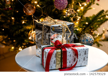 Stacked Christmas Gifts with Red Ribbon by Glowing Tree. Festive Christmas gifts wrapped in gold and white paper, one tied with a red ribbon and bow, stacked on a white table before a glowing Christma Stacked Christmas Gifts with Red Ribbon by Glowing Tree. Festive Christmas gifts wrapped in gold and white paper, one tied with a red ribbon and bow, stacked on a white table before a glowing Christma 142579329