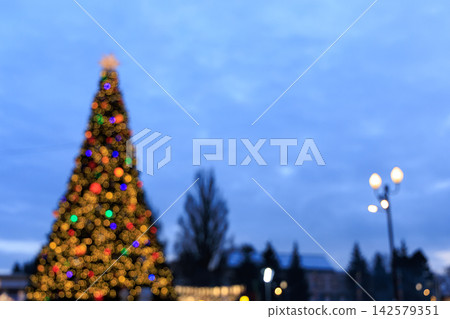 Holiday Christmas tree stands in a public square during evening with lights glowing under a darkening sky Holiday Christmas tree stands in a public square during evening with lights glowing under a darkening sky 142579351
