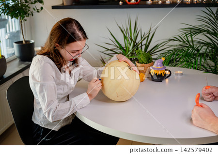 Daughter Decorating Pumpkin at Festive Halloween Table. A daughter with glasses decorates a pumpkin at a bright table with a witch-hat ornament, houseplants and candles. A cozy autumn scene of Hallowe Daughter Decorating Pumpkin at Festive Halloween Table. A daughter with glasses decorates a pumpkin at a bright table with a witch-hat ornament, houseplants and candles. A cozy autumn scene of Hallowe 142579402