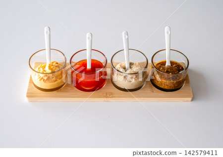 Four glass sauce bowls with ceramic spoons on wooden base. Four glass sauce bowls with white ceramic spoons on natural wooden bamboo base, colorful condiment serving set for kitchen styling and culina Four glass sauce bowls with ceramic spoons on wooden base. Four glass sauce bowls with white ceramic spoons on natural wooden bamboo base, colorful condiment serving set for kitchen styling and culina 142579411