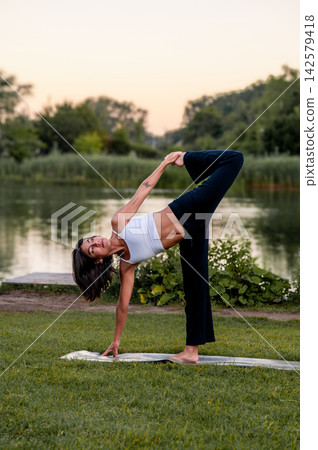 Woman in Standing Forward Fold in a Green Park. A young Hispanic woman practises a standing forward fold in a green park beside a calm lake at sunset, promoting balance, calm and inner peace Woman in Standing Forward Fold in a Green Park. A young Hispanic woman practises a standing forward fold in a green park beside a calm lake at sunset, promoting balance, calm and inner peace 142579418