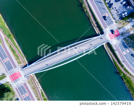 Aerial Top View of an Arched Bridge Over a Canal. Aerial top-down view of a white arched bridge crossing a green canal with roads, crosswalks and cars on both banks, a symmetrical drone view Aerial Top View of an Arched Bridge Over a Canal. Aerial top-down view of a white arched bridge crossing a green canal with roads, crosswalks and cars on both banks, a symmetrical drone view 142579419