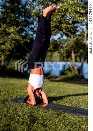 Fit Woman in Supported Headstand in the Park at Sunset. A young Hispanic woman practises a supported headstand surrounded by greenery in a park at golden hour, a moment of self care and active summer Fit Woman in Supported Headstand in the Park at Sunset. A young Hispanic woman practises a supported headstand surrounded by greenery in a park at golden hour, a moment of self care and active summer 142579420