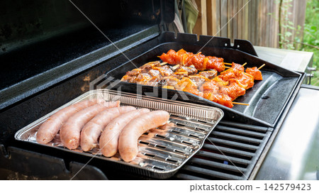Barbecue with Sausages and Marinated Skewers in Garden. Sausages in a foil tray and assorted marinated meat skewers grilling on an outdoor gas barbecue with green garden behind. A summer cookout scene 142579423
