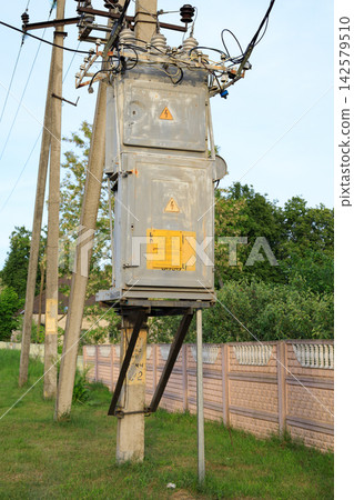 Old power transformer on a concrete pole near a fence. Rural electricity distribution infrastructure with high voltage cables Old power transformer on a concrete pole near a fence. Rural electricity distribution infrastructure with high voltage cables 142579510