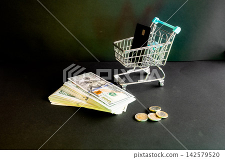Shopping Cart With a Bank Card Over Money. A miniature shopping cart holding a black bank card above a stack of dollar bills and coins on dark, a creative concept of online shopping and card payment. Shopping Cart With a Bank Card Over Money. A miniature shopping cart holding a black bank card above a stack of dollar bills and coins on dark, a creative concept of online shopping and card payment. 142579520