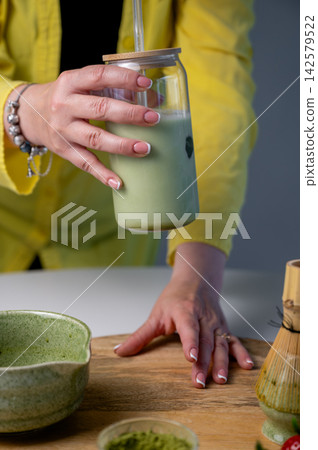 Woman's hands in Yellow Jacket Holding Matcha Latte Jar. Hands in yellow jacket gripping iced matcha latte jar with straw beside artisan tea preparation setup on wooden tray. Ceramic bowl and chasen Woman's hands in Yellow Jacket Holding Matcha Latte Jar. Hands in yellow jacket gripping iced matcha latte jar with straw beside artisan tea preparation setup on wooden tray. Ceramic bowl and chasen 142579522