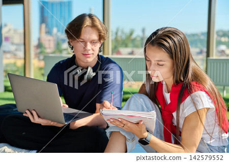 Two young university students sitting on floor studying using laptop and writing in notebook near window Two young university students sitting on floor studying using laptop and writing in notebook near window 142579552