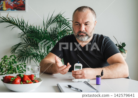 Mature Man Doing a Finger Blood Sugar Test. Mature man does a finger- blood sugar test with a glucometer at a table with a bowl of fresh strawberries, illustrating diabetes self-monitoring and a healt Mature Man Doing a Finger Blood Sugar Test. Mature man does a finger- blood sugar test with a glucometer at a table with a bowl of fresh strawberries, illustrating diabetes self-monitoring and a healt 142579641