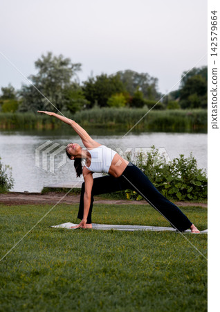 Fit Woman in Extended Side Angle Pose in a Green Park. A young Hispanic woman practises the extended side angle pose in a green park beside a calm lake at sunset, for stress relief and a healthy activ Fit Woman in Extended Side Angle Pose in a Green Park. A young Hispanic woman practises the extended side angle pose in a green park beside a calm lake at sunset, for stress relief and a healthy activ 142579664