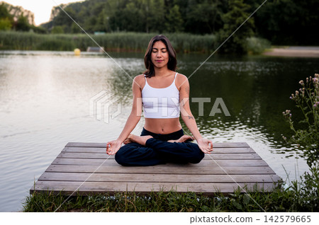 Young Woman in Seated Cross Legged Meditation in a Green Park. A young Hispanic woman practises a seated cross legged meditation in a green park beside a calm lake at sunset, nurturing wellness Young Woman in Seated Cross Legged Meditation in a Green Park. A young Hispanic woman practises a seated cross legged meditation in a green park beside a calm lake at sunset, nurturing wellness 142579665
