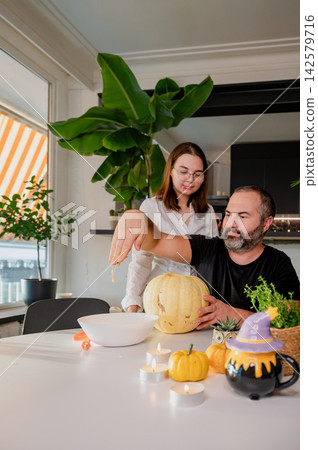 Father and Daughter at Table with Carved Pumpkin and Festive Decor. A father and his daughter clean a carved pale pumpkin at a bright table with mini pumpkins, candles and a witch-hat teapot in a cozy Father and Daughter at Table with Carved Pumpkin and Festive Decor. A father and his daughter clean a carved pale pumpkin at a bright table with mini pumpkins, candles and a witch-hat teapot in a cozy 142579716