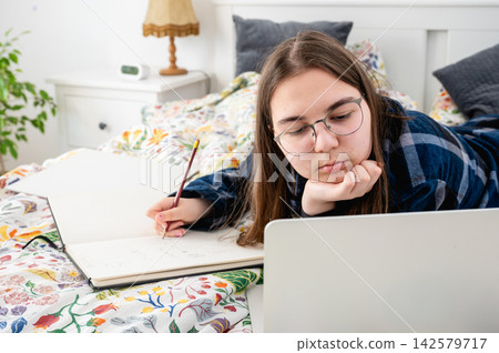 Teen Girl with Glasses Studying on Laptop in Cozy Evening Light. Teenage girl wearing glasses concentrating on homework with laptop and notebook in cozy evening bedroom. Warm lamp glow and floral bed Teen Girl with Glasses Studying on Laptop in Cozy Evening Light. Teenage girl wearing glasses concentrating on homework with laptop and notebook in cozy evening bedroom. Warm lamp glow and floral bed 142579717