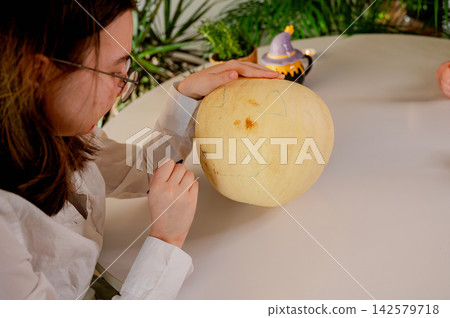 Daughter Carving Pumpkin with Tool for Halloween at Home, A daughter with glasses carves a pale pumpkin with a small tool at a white table decorated for Halloween. A cozy autumn scene of festive holid Daughter Carving Pumpkin with Tool for Halloween at Home, A daughter with glasses carves a pale pumpkin with a small tool at a white table decorated for Halloween. A cozy autumn scene of festive holid 142579718