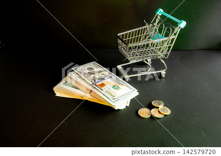 Miniature Cart With a Bank Card and Dollars. A miniature shopping cart with a black bank card beside US dollar bills and coins on a dark background, a conceptual image of online shopping and card paym Miniature Cart With a Bank Card and Dollars. A miniature shopping cart with a black bank card beside US dollar bills and coins on a dark background, a conceptual image of online shopping and card paym 142579720