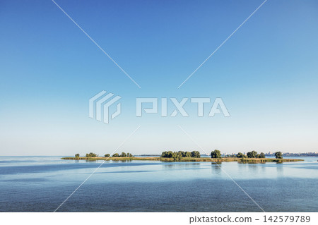 Small islands emerging from the calm water of a river under blue sky Small islands emerging from the calm water of a river under blue sky 142579789