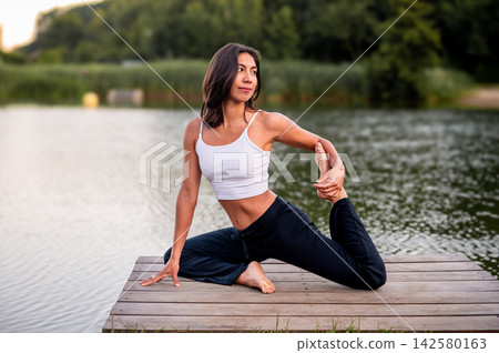 Hispanic Woman in Seated One Legged Pose in a Green Park. A young Hispanic woman practises a seated one legged pose in a green park beside a calm lake at sunset, embracing an active and mindful lifest 142580163