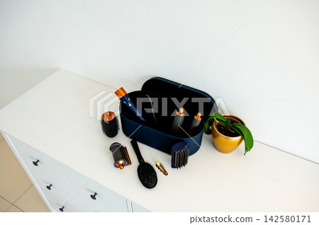 Open hair styler case with round brush and clip on dresser. Open navy hair styler case showing tools inside with round barrel brush and hair clip on white dresser, beauty technology and modern hair ca Open hair styler case with round brush and clip on dresser. Open navy hair styler case showing tools inside with round barrel brush and hair clip on white dresser, beauty technology and modern hair ca 142580171