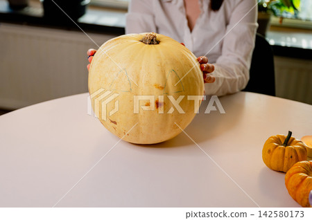 Pumpkin with Drawn Face Outline on Halloween Table. A pale pumpkin marked with a drawn face design held on a round white table beside mini pumpkins, ready for Halloween carving. A cozy autumn scene Pumpkin with Drawn Face Outline on Halloween Table. A pale pumpkin marked with a drawn face design held on a round white table beside mini pumpkins, ready for Halloween carving. A cozy autumn scene 142580173