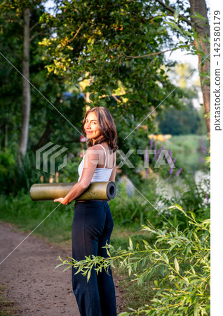 Fit Woman in Woman Carrying a Yoga Mat in a Summer Park. A young Hispanic woman carries a rolled yoga mat in a lush green park in the evening light on her way to an outdoor yoga session, embracing 142580179