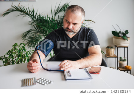 Mature Man Reading Blood Pressure Result at Home. Mature man checks his blood pressure result with a home arm-cuff monitor at a table with blister packs of pills, illustrating hypertension self-monito Mature Man Reading Blood Pressure Result at Home. Mature man checks his blood pressure result with a home arm-cuff monitor at a table with blister packs of pills, illustrating hypertension self-monito 142580184