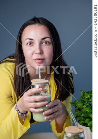 Relaxed Woman Enjoying Matcha Latte at Home, Summer. Relaxed woman enjoying matcha latte through straw at home in cozy summer setting with potted plant. Glass jar with green beverage and warm natural  142580191