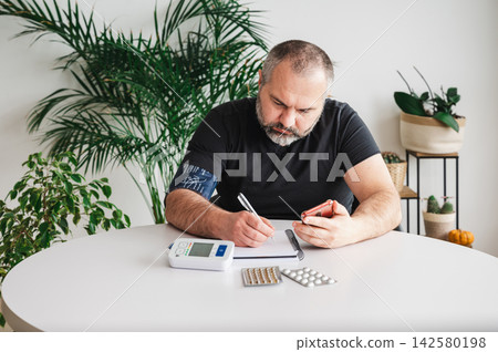 Mature Man Tracking Blood Pressure in a Notebook at Home. Mature man tracks his blood pressure in a notebook beside a home monitor and blister packs of pills at a table, illustrating health tracking Mature Man Tracking Blood Pressure in a Notebook at Home. Mature man tracks his blood pressure in a notebook beside a home monitor and blister packs of pills at a table, illustrating health tracking 142580198
